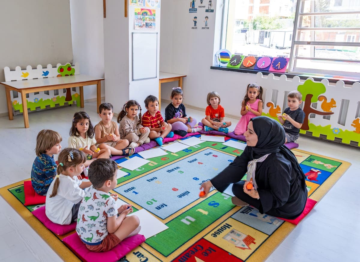Halının üzerinde öğretmenleriyle çember olmuş, oyun tahtasıyla çalışan çocuklar